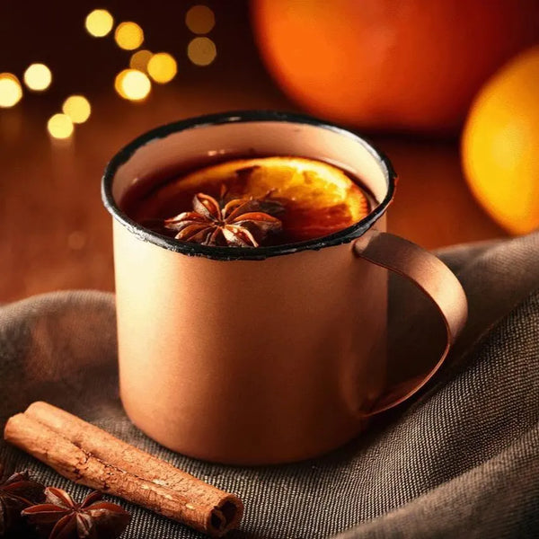 Copper mug with hot beverage and spices on a dark fabric background with blurred lights and pumpkins.