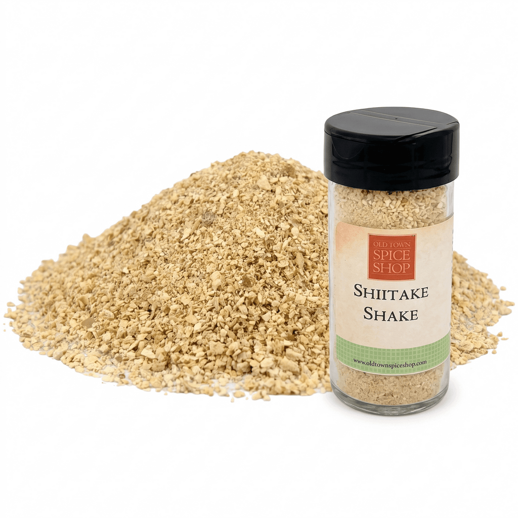 A clear plastic spice jar labeled 'Shiitake Shake' by The Awesome Spice Shop, standing next to a large mound of the light tan ground spice blend on a white background.