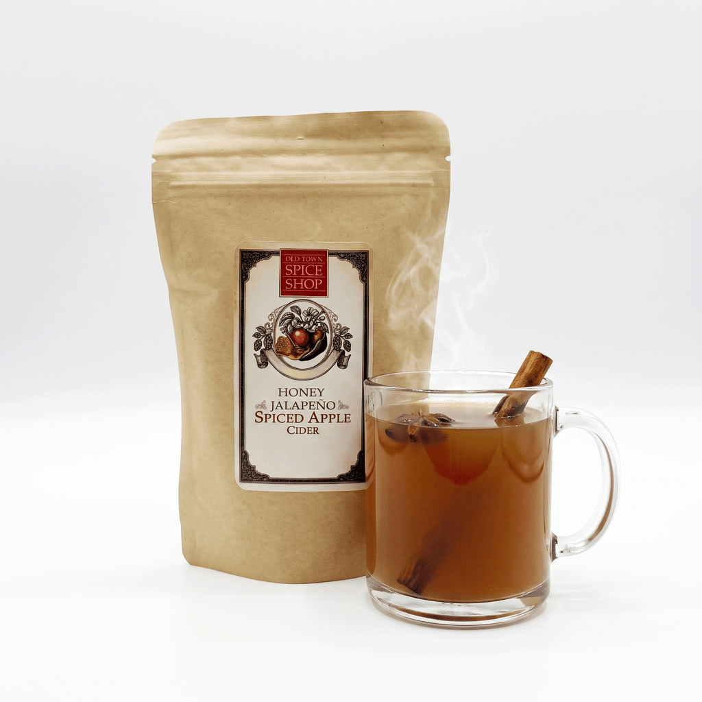 A kraft paper stand-up pouch labeled Spice Shop Honey Jalapeño and Spiced Apple Cider stands next to a clear glass mug filled with amber-brown spiced cider, cinnamon sticks, and visible steam, on a clean white background.