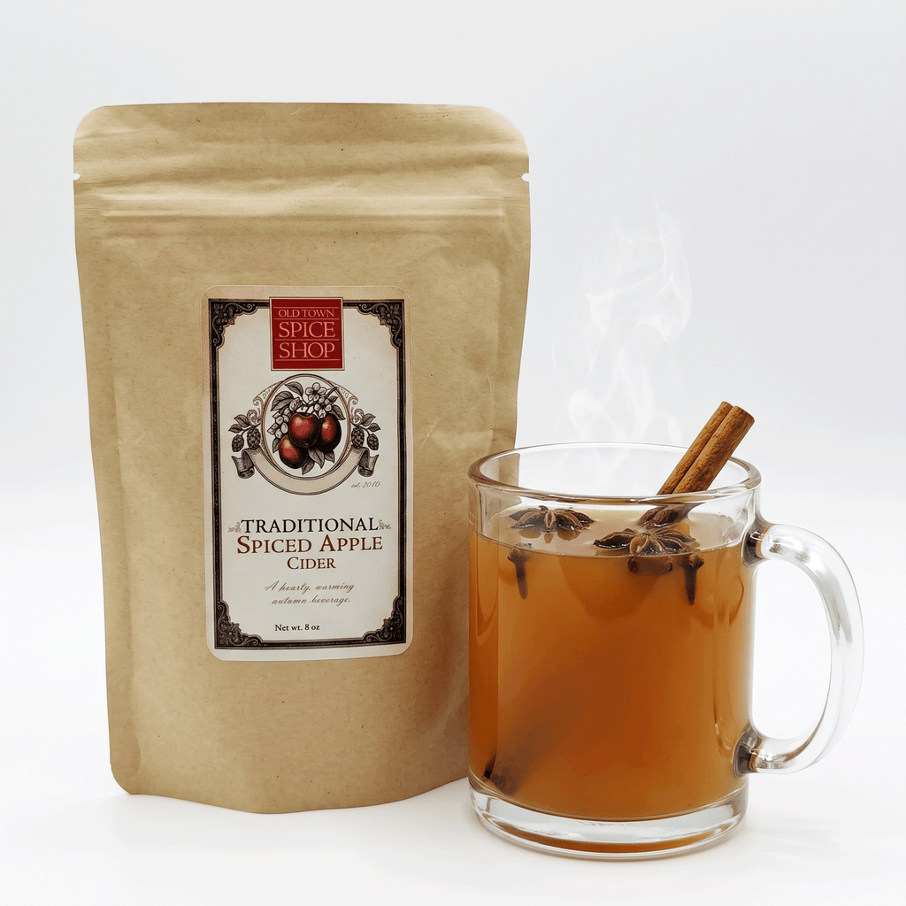 A kraft paper pouch of Old Town Spice Shop Traditional Spiced Apple Cider stands next to a clear glass mug filled with amber-colored cider, topped with visible steam and garnished with a cinnamon stick and star anise, on a clean white background.
