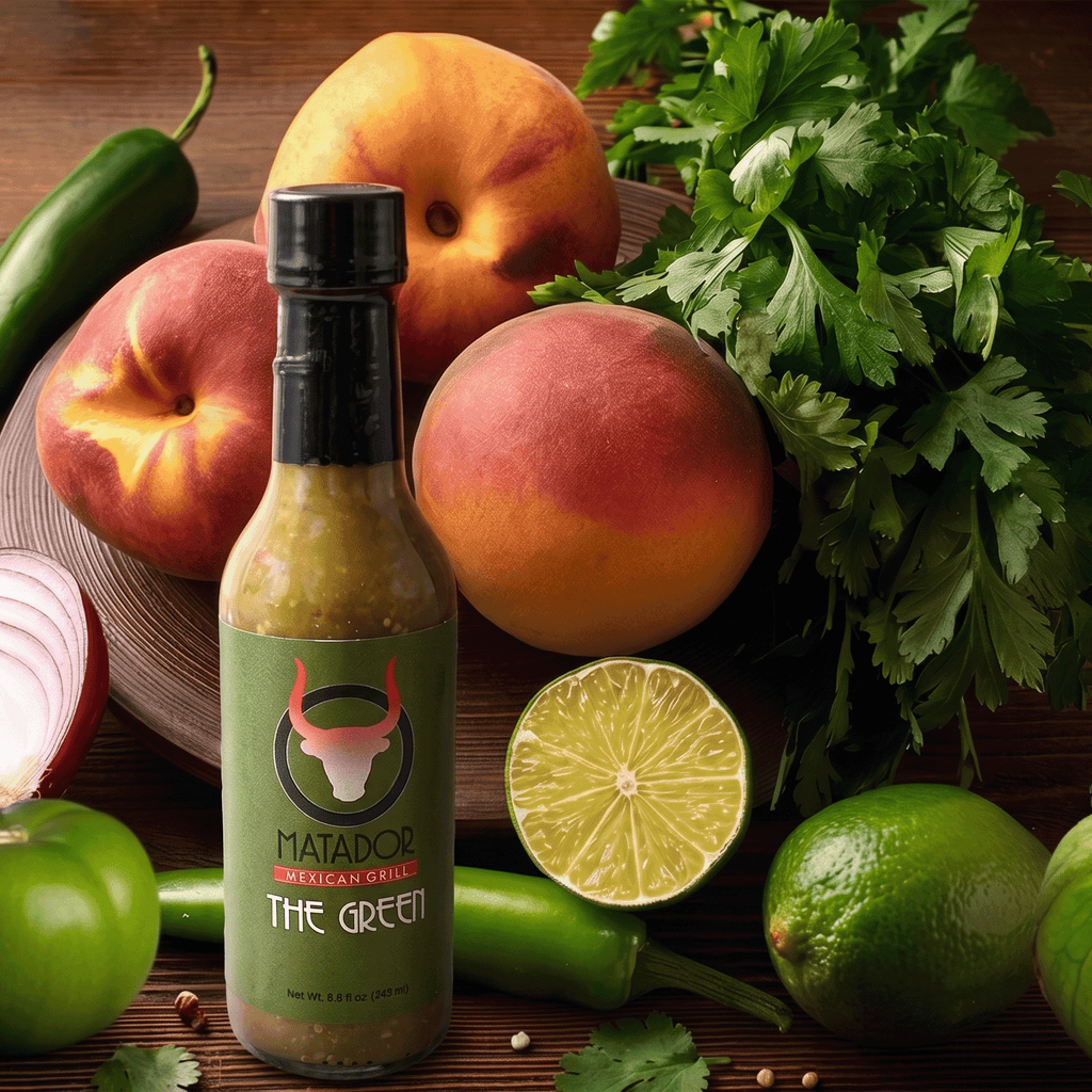 Bottle of Matador Mexican Grill The Green hot sauce surrounded by peaches, limes, and herbs on a wooden surface.