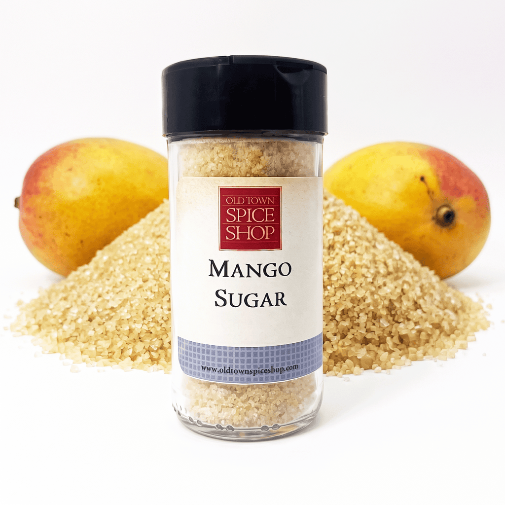 A clear plastic jar of Old Town Spice Shop Mango Sugar with a black lid and white label, set against a white background with loose brown sugar spilled in mounds and two whole mangoes behind it.